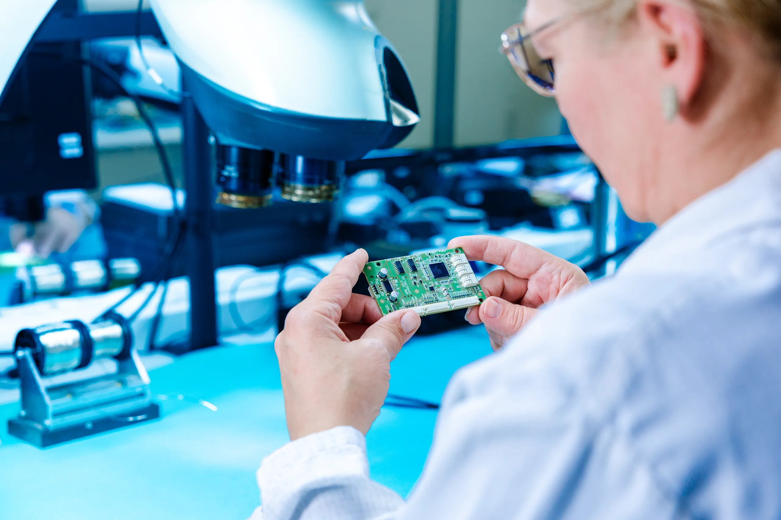 Mitarbeiterin kontrolliert eine bestückte Leiterplatte am Arbeitsplatz mit optischem Prüfgerät